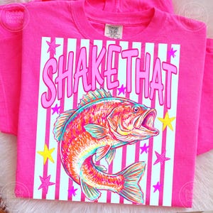 Puede incluir: Una camiseta rosa brillante con la frase "SHAKE THAT" en letras rosas. La camiseta presenta una colorida ilustraci&oacute;n de un pez con la boca abierta, sobre un fondo a rayas con acentos de estrellas. La camiseta est&aacute; hecha de un material suave.