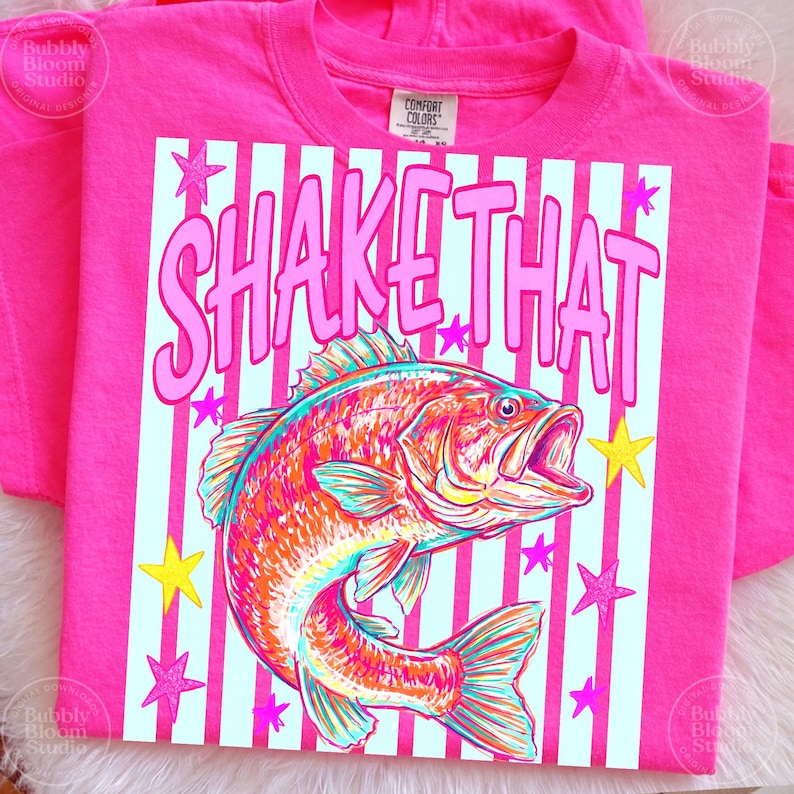 Puede incluir: Una camiseta rosa brillante con la frase "SHAKE THAT" en letras rosas. La camiseta presenta una colorida ilustraci&oacute;n de un pez con la boca abierta, sobre un fondo a rayas con acentos de estrellas. La camiseta est&aacute; hecha de un material suave.
