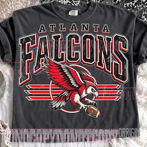 May include: A charcoal gray short-sleeve t-shirt featuring a distressed graphic of the Atlanta Falcons. The design includes the team name in large black and red lettering above a red and white falcon illustration holding a brown football.