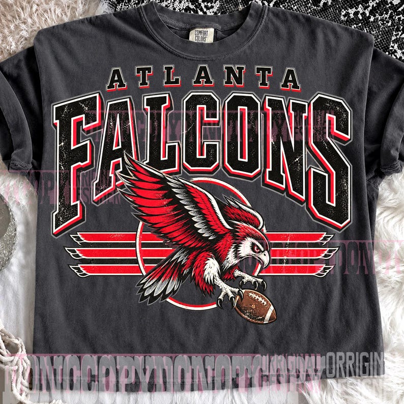 May include: A charcoal gray short-sleeve t-shirt featuring a distressed graphic of the Atlanta Falcons. The design includes the team name in large black and red lettering above a red and white falcon illustration holding a brown football.