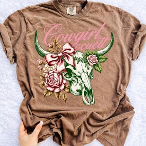 Peut inclure: T-shirt marron avec un motif de cr&acirc;ne de vache orn&eacute; d'un n&oelig;ud rose et de roses. Le texte "Cowgirl Society" est arqu&eacute; au-dessus du cr&acirc;ne en rose. Le t-shirt a des manches courtes et un col rond.