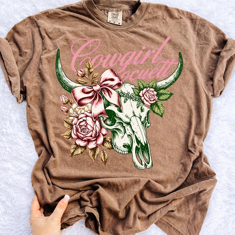 Peut inclure: T-shirt marron avec un motif de cr&acirc;ne de vache orn&eacute; d'un n&oelig;ud rose et de roses. Le texte "Cowgirl Society" est arqu&eacute; au-dessus du cr&acirc;ne en rose. Le t-shirt a des manches courtes et un col rond.