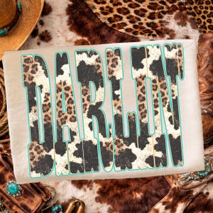 May include: A beige t-shirt with the word "DARLIN" in a bold, stylized font. The letters feature a mix of leopard and cow print patterns, outlined in a turquoise color. The shirt is surrounded by western-themed accessories.