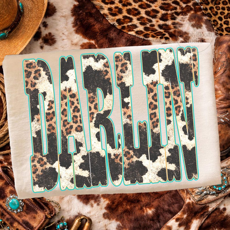 May include: A beige t-shirt with the word "DARLIN" in a bold, stylized font. The letters feature a mix of leopard and cow print patterns, outlined in a turquoise color. The shirt is surrounded by western-themed accessories.