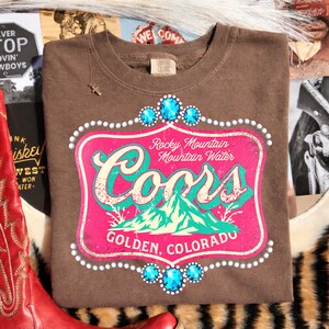 Peut inclure: T-shirt marron avec un motif r&eacute;tro. Le graphisme comprend le texte "Coors" et "Rocky Mountain Mountain Water" en rose et turquoise. Le design comporte des ornements de bijoux.