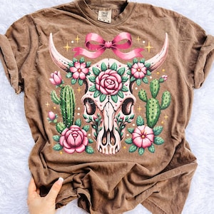 Peut inclure: T-shirt marron avec un motif de cr&acirc;ne de vache orn&eacute; de roses roses, d'un n&oelig;ud rose et de cactus. Le design a un aspect vintage et vieilli. Le t-shirt est fait d'une mati&egrave;re douce.