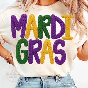May include: Cream-colored t-shirt with the words "MARDI GRAS" in large, textured letters. The letters are in purple, yellow, and green, with a knitted appearance. The shirt is worn by a person.