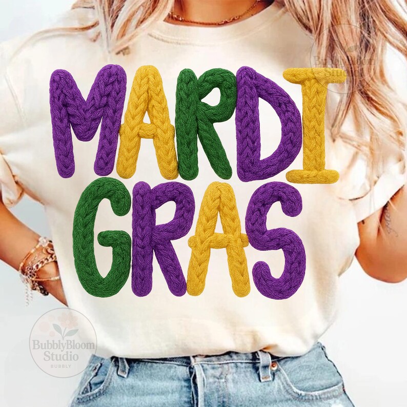 May include: Cream-colored t-shirt with the words "MARDI GRAS" in large, textured letters. The letters are in purple, yellow, and green, with a knitted appearance. The shirt is worn by a person.