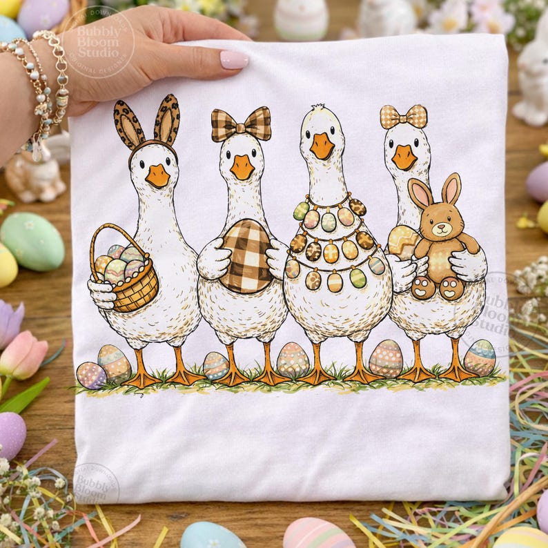 May include: White t-shirt featuring four geese dressed for Easter. Each goose holds an Easter egg or a basket of eggs, and wears a bow, bunny ears, or a necklace of eggs. The design includes colorful Easter eggs and a stuffed bunny.