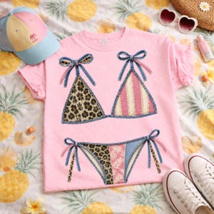 May include: Pink t-shirt featuring a bikini design. The bikini top has leopard print and pink and white stripes. The bikini bottom has leopard print, a floral print, and blue denim accents. A blue and yellow baseball cap is visible.