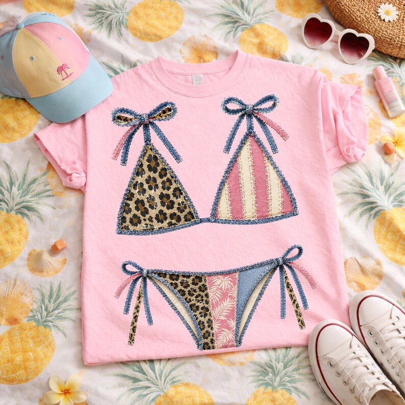 May include: Pink t-shirt featuring a bikini design. The bikini top has leopard print and pink and white stripes. The bikini bottom has leopard print, a floral print, and blue denim accents. A blue and yellow baseball cap is visible.