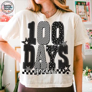 May include: Cream-colored t-shirt with a graphic celebrating 100 days of school. The design features the number "100" with a checkered pattern, the words "DAYS of school", and star accents. The shirt has a relaxed fit.
