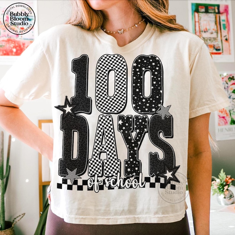 May include: Cream-colored t-shirt with a graphic celebrating 100 days of school. The design features the number "100" with a checkered pattern, the words "DAYS of school", and star accents. The shirt has a relaxed fit.