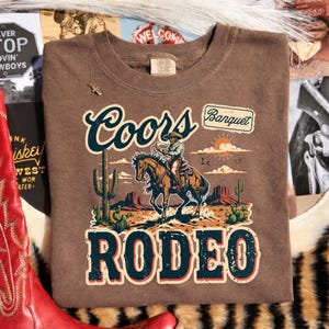 Peut inclure: T-shirt marron avec un motif repr&eacute;sentant un cow-boy &agrave; cheval, les mots "Coors Banquet Rodeo" et un paysage d&eacute;sertique. Le t-shirt a un col rond et une &eacute;tiquette de la marque Comfort Colors.