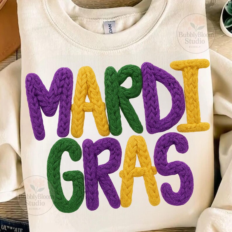 May include: Cream-colored sweatshirt with the words "MARDI GRAS" in large, textured, purple, green, and yellow letters. The letters have a knitted appearance, creating a festive and eye-catching design.