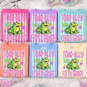 Könnte beinhalten: Sechs farbenfrohe T-Shirts mit dem Text "TOAD-ALLY Into" und einer Klassenstufe, mit einem Cartoon-Frosch und Sternen. Die Shirts sind in Pink, Lila, Hellblau, Orange, Blau und Gelb.