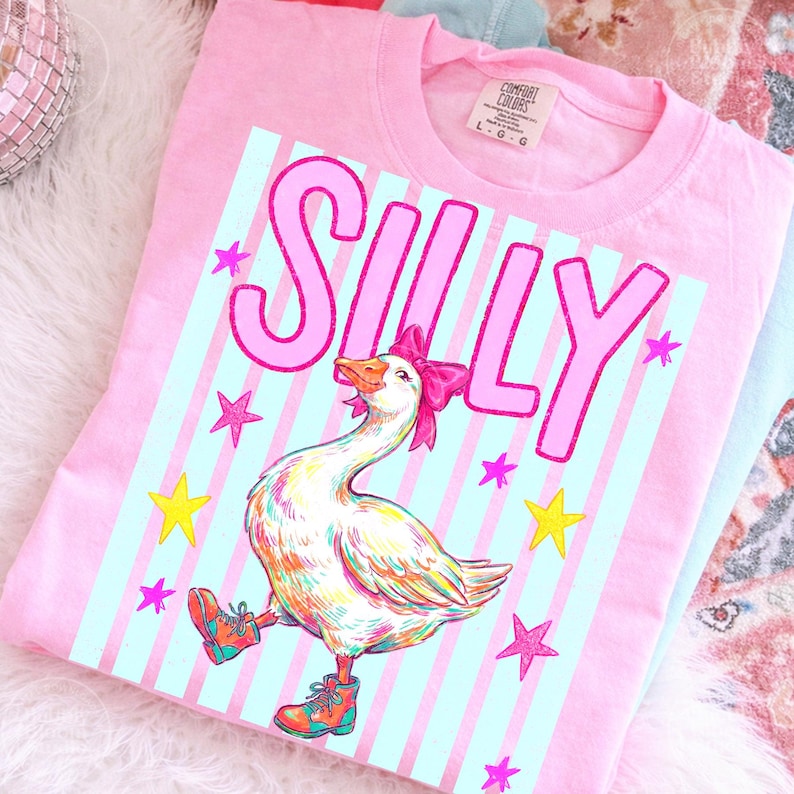 Puede incluir: Camiseta rosa Comfort Colors con un gr&aacute;fico de un ganso con un lazo rosa y botas, con la palabra "SILLY" encima. El ganso est&aacute; sobre un fondo a rayas azul claro con estrellas rosas y amarillas.