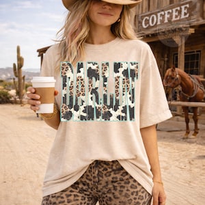 May include: A beige t-shirt with the word "DARLIN" in a turquoise outline, filled with a cow print and leopard print design. The person is wearing leopard print pants and a cowboy hat, holding a coffee cup.