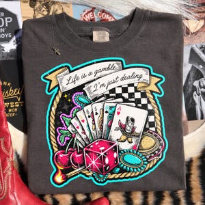 Peut inclure: T-shirt gris fonc&eacute; avec un motif color&eacute; de cartes &agrave; jouer, de d&eacute;s et de cerises. Le texte "Life is a gamble, I'm just dealing" est affich&eacute; sur une banni&egrave;re. Col rond et manches courtes.