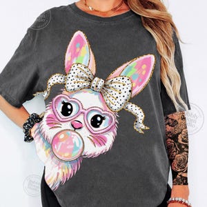 May include: Dark gray t-shirt featuring a cartoon bunny with heart-shaped glasses, a polka-dot bow, and a bubblegum bubble. The bunny's ears and bubble are rainbow-colored. The shirt has a vintage, washed look.