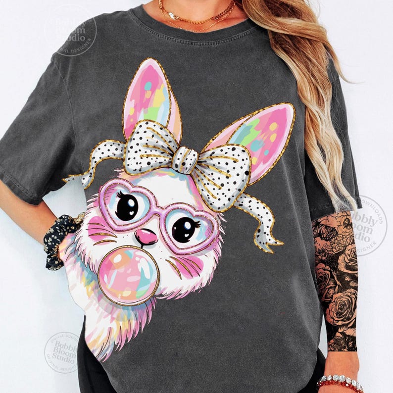 May include: Dark gray t-shirt featuring a cartoon bunny with heart-shaped glasses, a polka-dot bow, and a bubblegum bubble. The bunny's ears and bubble are rainbow-colored. The shirt has a vintage, washed look.
