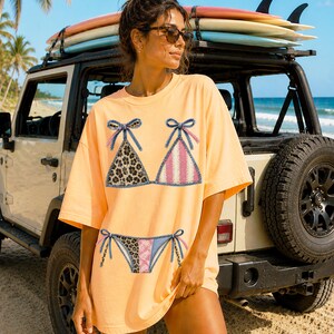 May include: A light orange t-shirt featuring a bikini design. The bikini top has a leopard print and pink stripes, with blue denim-style trim and bows. The matching bikini bottom is also visible. The shirt is worn near a beach with a jeep and surfboards.