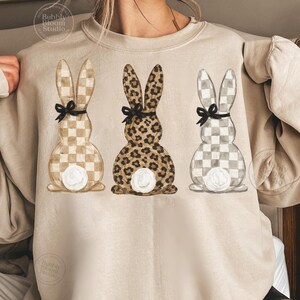 May include: Beige sweatshirt featuring three bunny designs. The bunnies have checkered and leopard print patterns, black bows, and white fluffy tails. The sweatshirt has a crew neck and long sleeves.