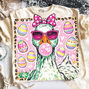 May include: Cream-colored t-shirt featuring a whimsical graphic of a goose wearing pink sunglasses and a bow, blowing a bubblegum bubble. The design includes Easter eggs and a leopard print border, perfect for Easter.