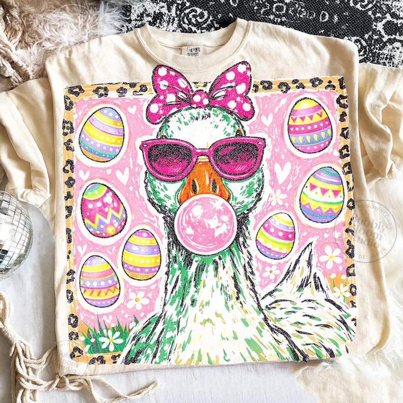 May include: Cream-colored t-shirt featuring a whimsical graphic of a goose wearing pink sunglasses and a bow, blowing a bubblegum bubble. The design includes Easter eggs and a leopard print border, perfect for Easter.