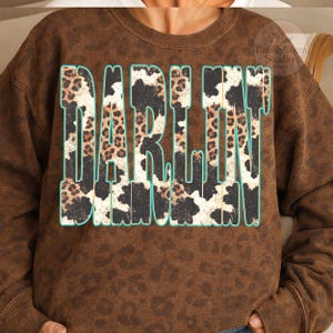 May include: Brown sweatshirt with the word "DARLIN" in large, bold letters. The letters have a leopard print and cow print design with a teal outline. The sweatshirt has a speckled pattern.