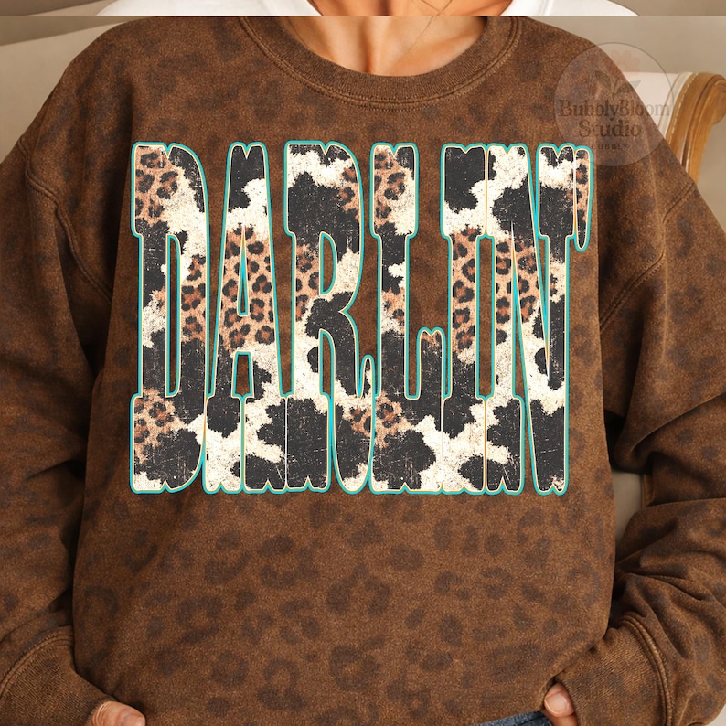 May include: Brown sweatshirt with the word "DARLIN" in large, bold letters. The letters have a leopard print and cow print design with a teal outline. The sweatshirt has a speckled pattern.