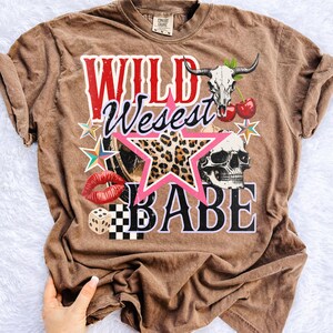 Peut inclure: T-shirt marron avec un motif graphique avec les mots "WILD West BABE". Le motif comprend un cr&acirc;ne, une &eacute;toile, des l&egrave;vres, des d&eacute;s et une &eacute;toile &agrave; imprim&eacute; l&eacute;opard. Le t-shirt a un aspect vintage et us&eacute;.