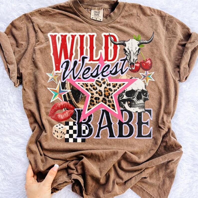 Peut inclure: T-shirt marron avec un motif graphique avec les mots "WILD West BABE". Le motif comprend un cr&acirc;ne, une &eacute;toile, des l&egrave;vres, des d&eacute;s et une &eacute;toile &agrave; imprim&eacute; l&eacute;opard. Le t-shirt a un aspect vintage et us&eacute;.