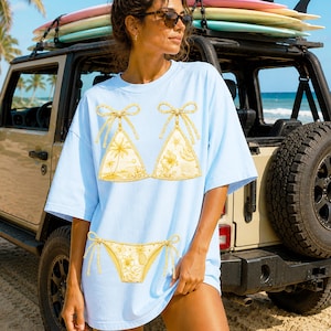 May include: Light blue t-shirt featuring a yellow bikini design. The bikini top has a bow and tropical floral and palm tree patterns. The matching bikini bottom is also yellow with a floral and pineapple pattern. A Jeep and surfboards are in the background.