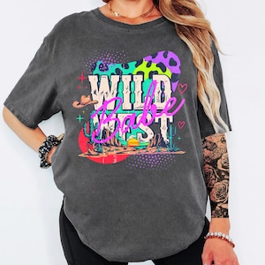 May include: Dark gray t-shirt with a colorful graphic that says "Wild Babe West." The design includes a cowboy hat, cactus, and a desert landscape with a sunset. The graphic has purple, green, pink, and orange accents.
