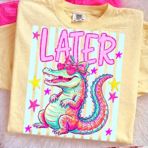 Puede incluir: Camiseta amarilla clara con un dise&ntilde;o de un caim&aacute;n colorido con gafas de sol en forma de coraz&oacute;n y un lazo. La palabra "LATER" est&aacute; impresa en rosa sobre el caim&aacute;n. Estrellas rosas y amarillas adornan el dise&ntilde;o.