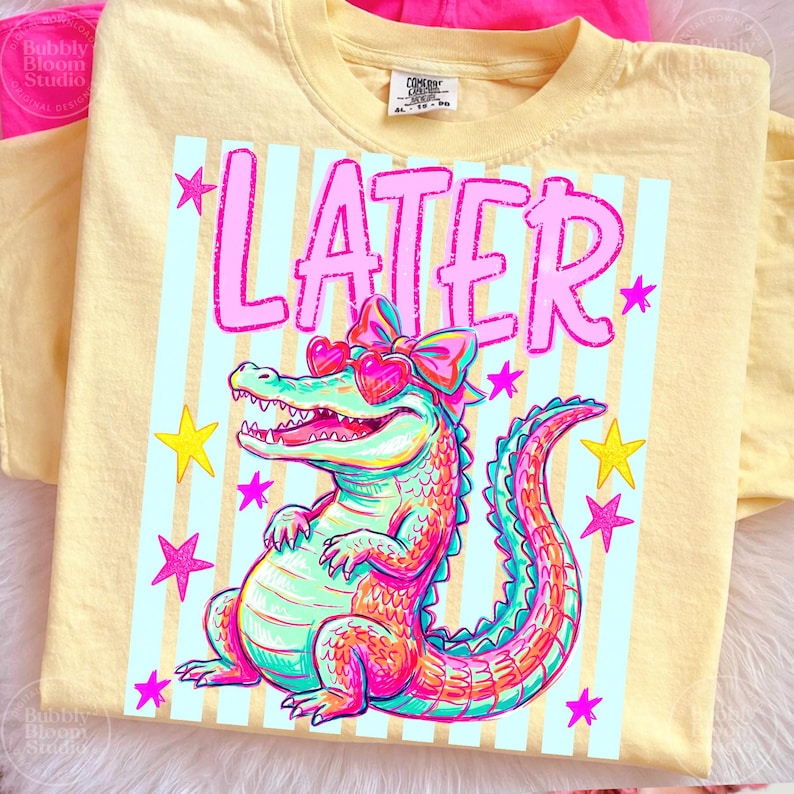 Puede incluir: Camiseta amarilla clara con un dise&ntilde;o de un caim&aacute;n colorido con gafas de sol en forma de coraz&oacute;n y un lazo. La palabra "LATER" est&aacute; impresa en rosa sobre el caim&aacute;n. Estrellas rosas y amarillas adornan el dise&ntilde;o.