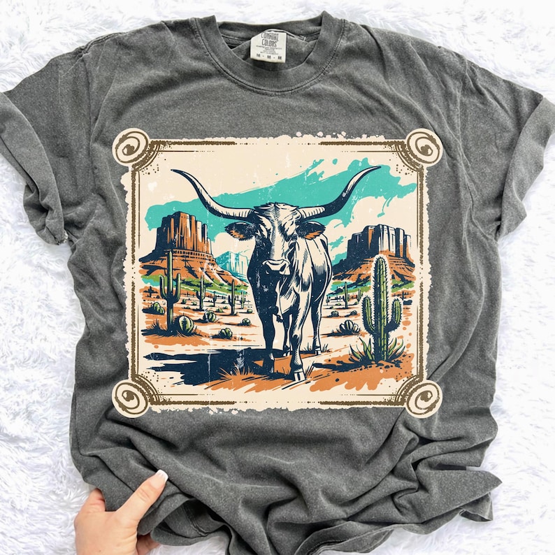 Peut inclure: T-shirt gris fonc&eacute; avec un motif de longhorn dans un paysage d&eacute;sertique. Le motif comprend des cactus, des mesas et un ciel turquoise, encadr&eacute; par une bordure d&eacute;corative. Le t-shirt est un v&ecirc;tement d&eacute;contract&eacute; et confortable.