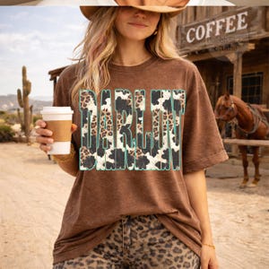May include: A brown t-shirt with the word "DARLIN" in a turquoise and cow print design. The person in the image is wearing a hat and leopard print pants, holding a coffee cup. The background includes a coffee shop sign and a horse.