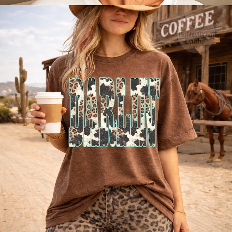 May include: A brown t-shirt with the word "DARLIN" in a turquoise and cow print design. The person in the image is wearing a hat and leopard print pants, holding a coffee cup. The background includes a coffee shop sign and a horse.