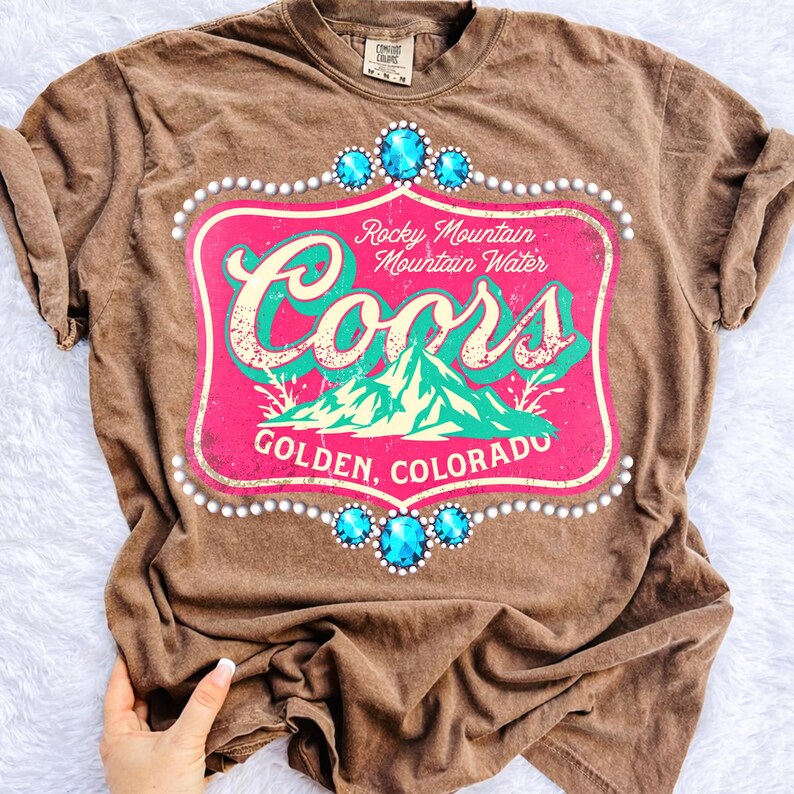 Peut inclure: T-shirt marron avec un motif vieilli. Le motif comprend le mot "Coors" en turquoise, un fond rose et des accents turquoise. Le motif comprend &eacute;galement le texte "Rocky Mountain Mountain Water" et "Golden, Colorado."
