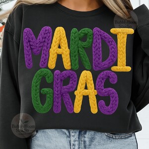 May include: Black sweatshirt with the words "MARDI GRAS" in large, textured letters. The letters are in purple, yellow, and green, with a knitted appearance. The sweatshirt is a casual style.