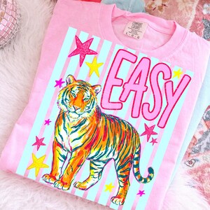 Puede incluir: Camiseta rosa con un gr&aacute;fico de tigre colorido, la palabra "EASY" y estrellas. El tigre es naranja, amarillo y rojo con rayas negras. La camiseta tiene un fondo a rayas azul claro.