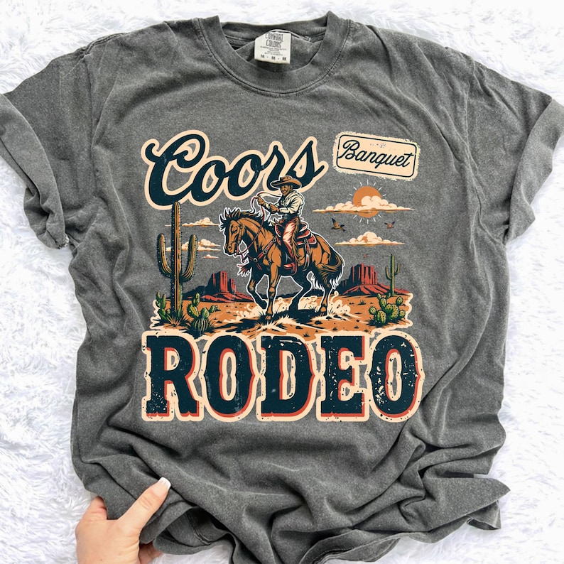 Peut inclure: T-shirt gris fonc&eacute; avec un motif sur le th&egrave;me du rod&eacute;o. Le design comprend un cow-boy &agrave; cheval, des cactus et les mots "Coors Banquet" et "Rodeo". Le t-shirt a un aspect vintage et us&eacute;.
