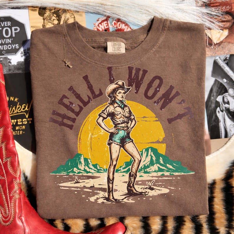 Peut inclure: T-shirt marron avec l'inscription "HELL I WON'T" et un graphisme de cowgirl en chapeau de cowboy, short et bottes, sur fond de soleil jaune et de montagnes. Le t-shirt est de Comfort Colors.