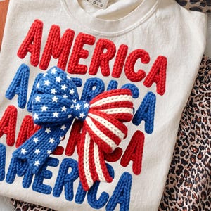 May include: Cream-colored sweatshirt with the word "AMERICA" in red, knitted letters. A blue bow with white stars and a red and white striped bow are centered on the shirt. The shirt is folded, with a leopard print pattern visible.