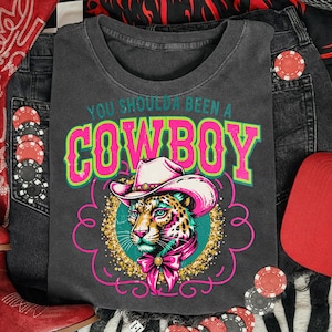 May include: Dark gray t-shirt with the text "YOU SHOULDA BEEN A COWBOY" in green and pink. The shirt features a leopard wearing a cowboy hat and a pink bow. The design is surrounded by a pink decorative border.