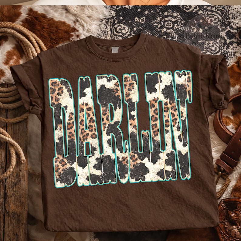 May include: A brown t-shirt with the word "DARLIN" in large, bold letters. The letters are filled with a leopard and cow print design, outlined in a teal color. The shirt has rolled-up sleeves.