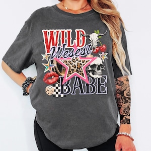 May include: Dark gray t-shirt with a graphic design featuring the text "WILD West BABE". The design includes a star with a leopard print, a skull, cherries, and a pair of red lips. The shirt has short sleeves.