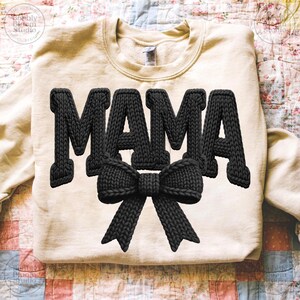 May include: A cream-colored sweatshirt with the word "MAMA" in large, black, knitted letters. A black knitted bow is centered below the word. The sweatshirt is a casual style, suitable for everyday wear.